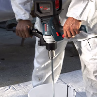 A person wearing a white protective suit holds a heavy-duty Bosch power mixer with both hands, lowering the mixing paddle into a white bucket.