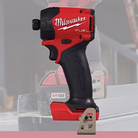 A cropped, side-view image of a red and black Milwaukee M18 Fuel cordless impact driver tool body. The tool features a textured black rubber grip, a red housing with the white Milwaukee logo, and a metal belt clip attached to the base where the battery slides in. A faded, desaturated image of the tool on a job site is visible in the background.