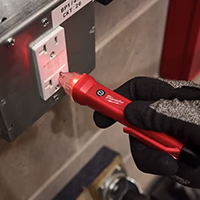 A close-up shot of a hand wearing a gray and black protective work glove, using a red Milwaukee non-contact voltage detector on a white GFCI outlet. The outlet is mounted in a metal junction box on a concrete wall with a label reading BP1/4/1 CKT 26. The tip of the voltage tester and an indicator on the outlet are both glowing red, indicating live electrical current.