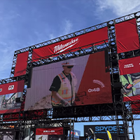 A massive outdoor scaffolding structure covered in red Milwaukee Tool branding banners. A large LED video screen in the center displays a construction worker in a hard hat and safety vest, with a countdown timer showing 0:48. Banners for M12, M18, MX Fuel, Hand Tools, and Accessories surround the screen under a bright blue sky.